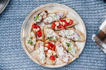 Bruschetta with tomatoes and shrimp salad on a metal plate, which stands on a knitted blue tablecloth. Nearby lies a checkered brown plaid. Top view