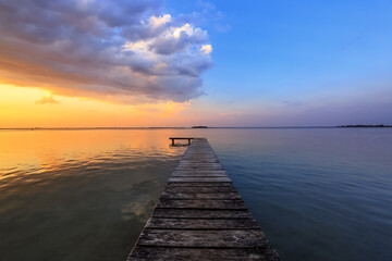 Fototapeta na wymiar Old wooden jetty, pier reveals views of the beautiful lake, blue sky with cloud. Sunrise enlightens the horizon with orange warm colors. Summer landscape. Free space for text.