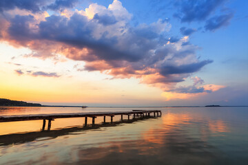 Old wooden jetty, pier reveals views of the beautiful lake, blue sky with cloud. Sunrise enlightens the horizon with orange warm colors. Summer landscape. Free space for text.
