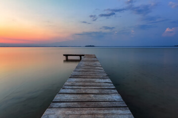 Fototapeta na wymiar Old wooden jetty, pier reveals views of the beautiful lake, blue sky with cloud. Sunrise enlightens the horizon with orange warm colors. Summer landscape. Free space for text.