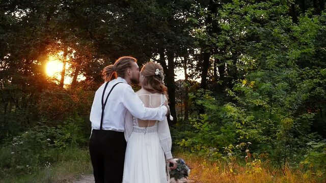 Loving young hipster couple walking in the park during a beautiful summer sunset. A guy with dreadlocks and a girl in a rustic wedding dress hold hands, hug and smile.
