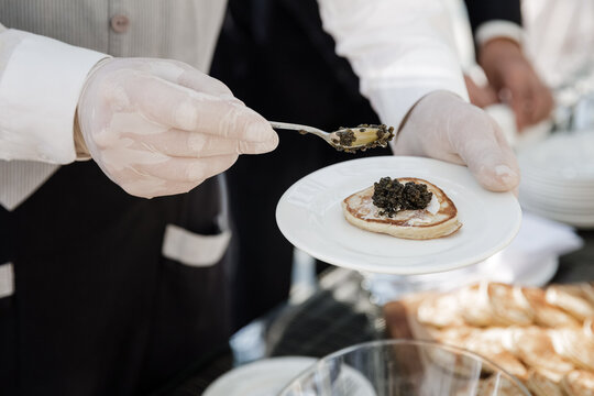 The waiter smears the pancakes with black caviar. A white plate in his hands. 