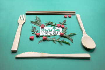 Close up of wood ecologic cutlery (fork, spoon, knife and chinese chopsticks), 
rocket salad and little red hearts. Eco vegan logo on green background. Concept of healthy and vegan alimentation. 
