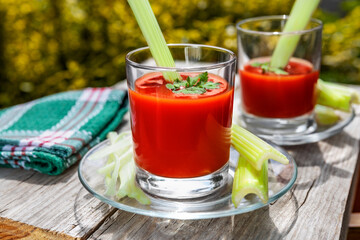 Two glasses of tomato juice with parsley and celery decorations, stand in the open air on wooden boards, the rays of the sun shine, shallow depth of field, selective focus. The concept of natural