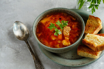 A bowl with Ukrainian soup-borscht decorated with a sprig of parsley on top, rad toasted bread, croutons. There is a spoon next to it. Close up..