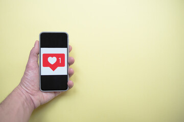 Close-up to a caucasic man hand holding a smartphone with heart icon on the screen. Yellow background. Concept of addiction to technology and social networks.