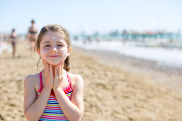 Cute little girl smiling and applying sunscreen on the face on the beach - summer vacation