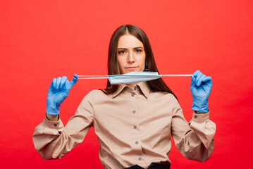 Cheerful and joyful young female playing with protective medical mask and blue gloves on red background. Close-up portrait. End of coronavirus quarantine, isolation. Covid-2019