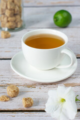 Breakfast. Tea in a white cup and saucer on a wooden white table. Brown sugar slices, two slices of gingerbread cookie, green lemon and white flower on the table. Vertical layout.