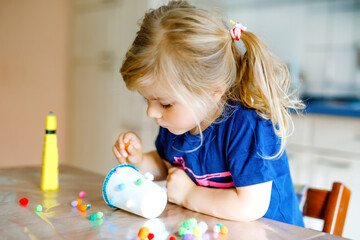 little toddler girl making craft lantern with paper cups, colorful pompoms and glue during pandemic...