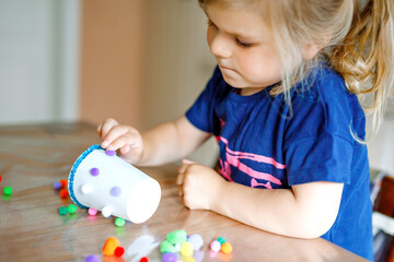little toddler girl making craft lantern with paper cups, colorful pompoms and glue during pandemic coronavirus quarantine disease. Happy creative child, homeschooling and home daycare with parents