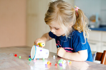 little toddler girl making craft lantern with paper cups, colorful pompoms and glue during pandemic...