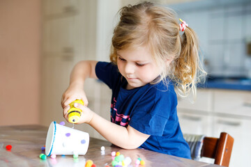 little toddler girl making craft lantern with paper cups, colorful pompoms and glue during pandemic coronavirus quarantine disease. Happy creative child, homeschooling and home daycare with parents