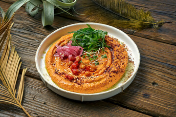 Classic oriental appetizer - baked peppers hummus with greens and arugula in a white plate on a wooden background