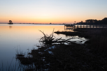 Brilliant sunrise in a salt marsh with a pier and dead tree silhouetted.