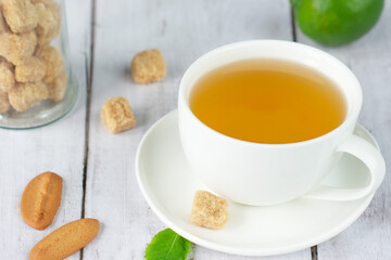 Breakfast. Tea in a white cup and saucer on a wooden white table. Brown sugar slices, two slices of gingerbread cookie, green lemon and white flower on the table. Horizontal layout.