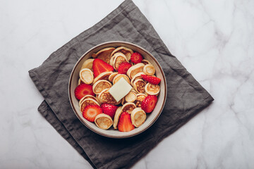 Trendy home breakfast with tiny cereal pancakes with piece of butter and fresh strawberries in the bowl. View from above.