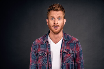 Redhead male with bristle dropping jaw as seeing incredible and amazing scenery, posing over gray background in casual shrit, gasping, staring thrilled and astonished at camera over grey wall