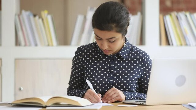 Focused smart indian ethnic girl college university high school student write notes essay homework assignment with textbook using laptop computer study in library prepare for exam sit at desk