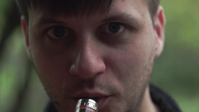 Pern exhales white smoke from electronic cigarette, vape into camera. Close-up beautiful eyes man looking camera in the country, against background grass trees, greens. Smoke fills entire video