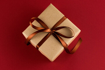A gift square box packed in craft paper and tied with a brown ribbon on a red background close-up.