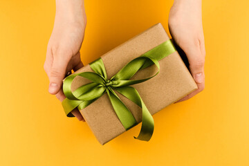 Female hands hold a gift box wrapped in craft paper and tied with a green ribbon on a yellow...