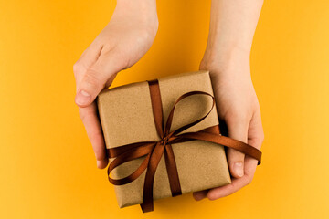 Female hands hold a gift box wrapped in craft paper and tied with a brown ribbon on a red background close-up.