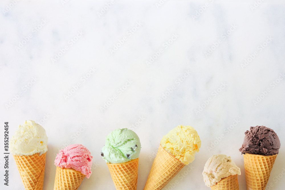 Poster Ice cream cone bottom border with a variety of flavors. Overhead view on a white marble background with copy space.