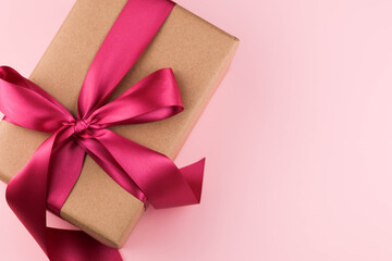 A gift box wrapped in craft paper and tied with a pink ribbon on a pink background close-up. Copy space