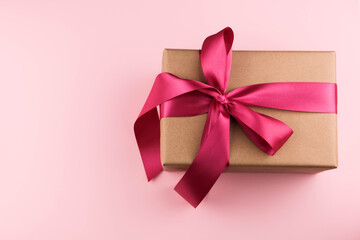A gift box wrapped in craft paper and tied with a pink ribbon on a pink background close-up. Copy space