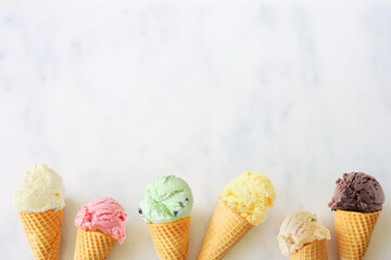 Ice cream cone bottom border with a variety of flavors. Overhead view on a white marble background with copy space.