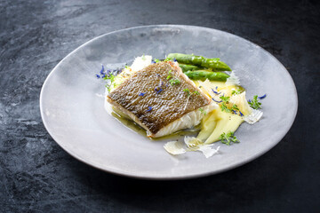 Traditionelles gebratenes Skrei Kabeljau Fisch Filet mit grünen Spargel Spitzen und Kartoffel Püree Creme in Parmesan Olivenöl Sauce als closeup auf einem Modern Design Teller mit Textfreiraum