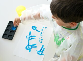 Boy painting with a brush and blue paint a white paper