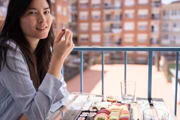 Asian girl eating sushi on the balcony sitting on a chair on the terrace in casual clothes