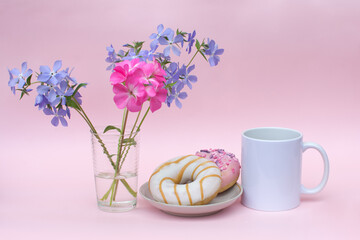 On a pink background-a white mug without logos. There are 2 delicious doughnuts on the plate. There are blue and pink flowers in the vase. Photos in pastel colors. Advertising a delicious Breakfast