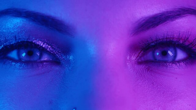 Extreme close up of human eyes iris under neon light 4K. Female with beautiful makeup, glitter shadows and false lashes. Womens green eyes contracting. Nightlife, night club concept.