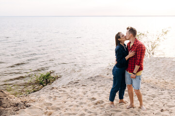 Young couple in love dressed in casual clothes barefoot stand on the sand near the sea and gently kissing and hugging