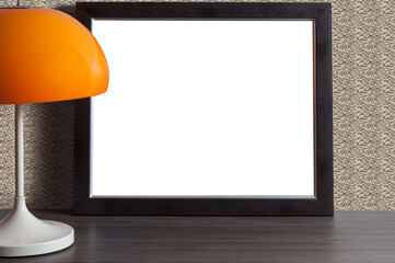 Empty black photo frame on a wooden table with a table lamp under an orange lampshade.
