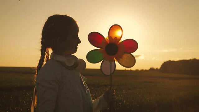 Cheerful girl are running in the meadow with wind turbines as a sign of freedom. Happy child holding wind toy running in green field, rejoicing life. Silhouette of kid girl holding wind toy.