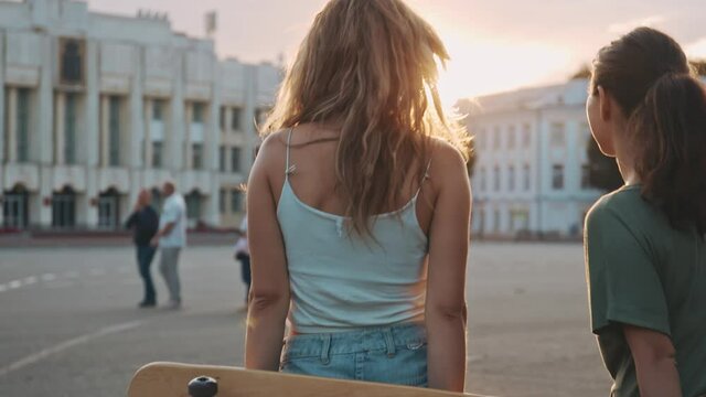 Concept of youth, active life and teen spirit. Two happy female friends spend time together skateboarding at sunset evening. Beautiful mixed-race girls having fun. Sport and friensdship lifestyle.