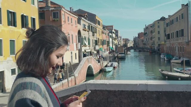 Beautiful asian woman tourist in red sweater enter in frame taking photo of the picturesque venetian canal with her smartphone from edge of the bridge. Charming woman spending her vacation in Venice.