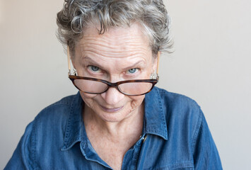 Close up of senior woman with blue eyes peering over glasses against neutral background (selective focus)