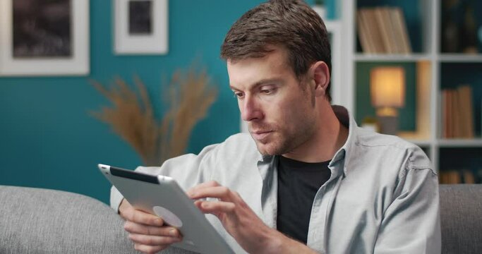 Close up of handsome man in domestic clothing sitting on couch and using digital tablet at home. Bearded guy enjoying free time with modern technology.