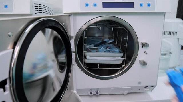 The laboratory technician in green uniform and white gloves quickly off-load the white autoclave with package dental tools after sterilization and disinfection in dental clinic. Unloading in fast