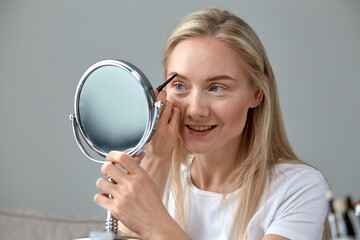 A young woman looks into a round mirror and draws her eyebrows with a pencil.