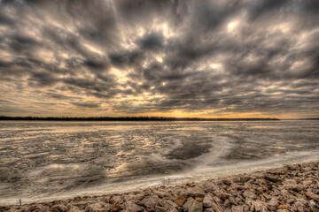 dramatic winter scene of a frozen Mississippi river stretching to the horizon along with cloudy sky, dark clouds blanket the sky with sunlight seeping through around edges, orange and yellow horizon 