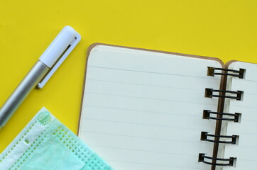 a blank notebook with a face mask, and a pen on yellow background.