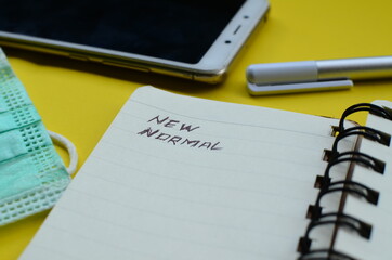 New Normal text on note pad with face mask ,cell phone, and pen on yellow background.