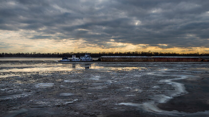 A towboat pushing a line of barges up the Mississippi river through ice, dark clouds above and sunlight coloring the sky yellow and orange in the distance, near Graftin Illinois