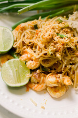 Traditional Shrimp Pad Thai served with lime wedges, noodles, and scallions with crushed peanuts. This delicious thai dish is served on a white plate.  This is a close-up food photo. 